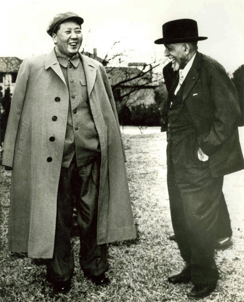 Mao Zedong in a long pale overcoat and flat cap, laughing, standing on grass beside W. E. B. Du Bois in a dark overcoat and hat, who is laughing back at him.